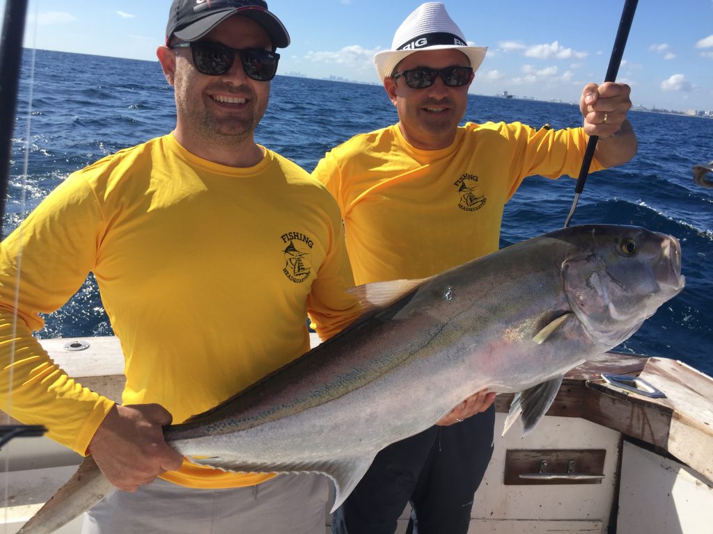 Couple guys in gold Fishing Headquarters shirts pose with a big amberjack they just caught off a sunken ship in Ft Lauderdale