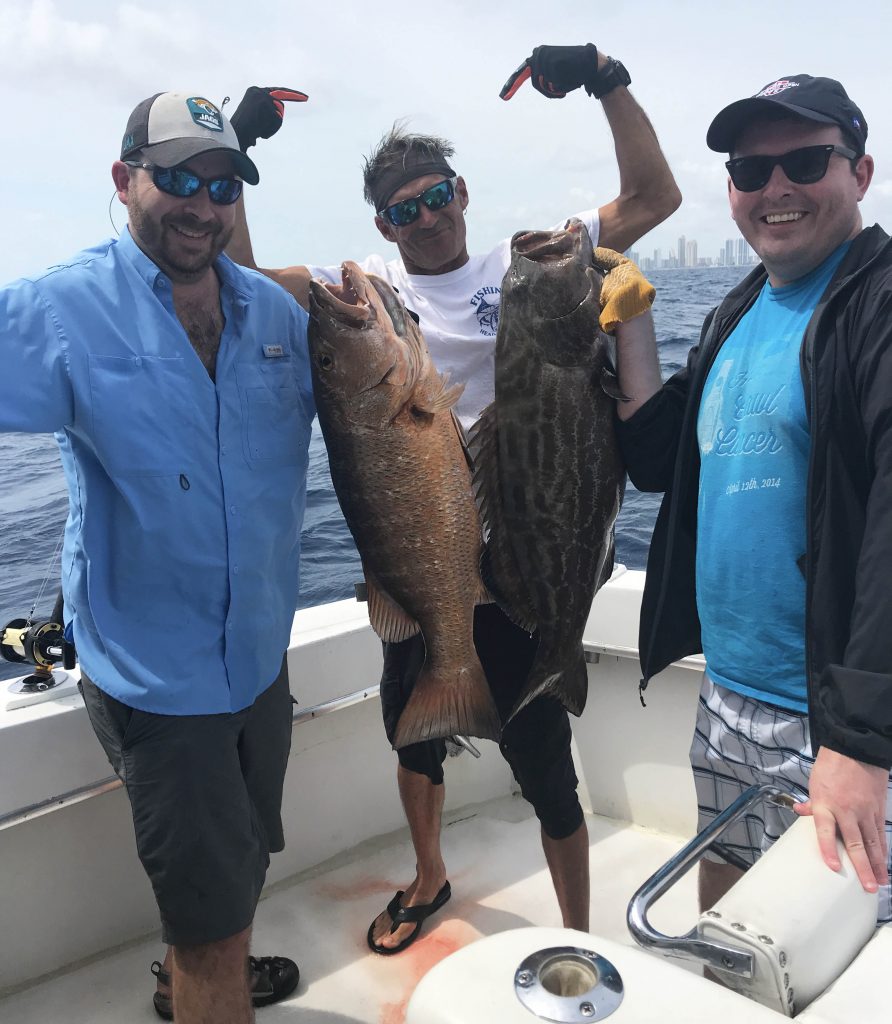 A couple of happy anglers holding a Cubera snapper and a black grouper aboard the boat with Dave in the background pointing at the fish.