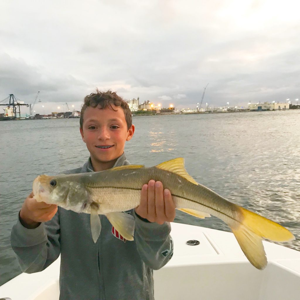 Adolescent boy holding a nice snook he just caught aboard the boat just around sunset on an inshore fishing charter with Port Everglades in the background.